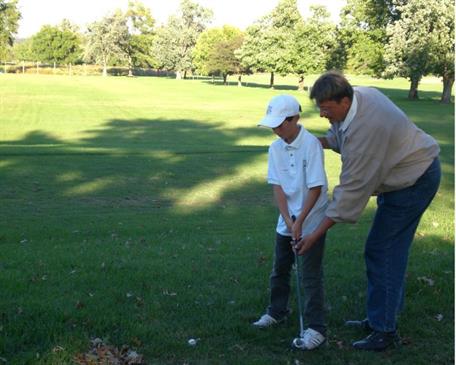 Junior playing golf in white polo/ Adult in neutral tone sweater is aiding